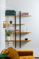 Solid wood floating shelf ÄIKE with two metal frames on a living room wall decorated with a plant, books and a candle