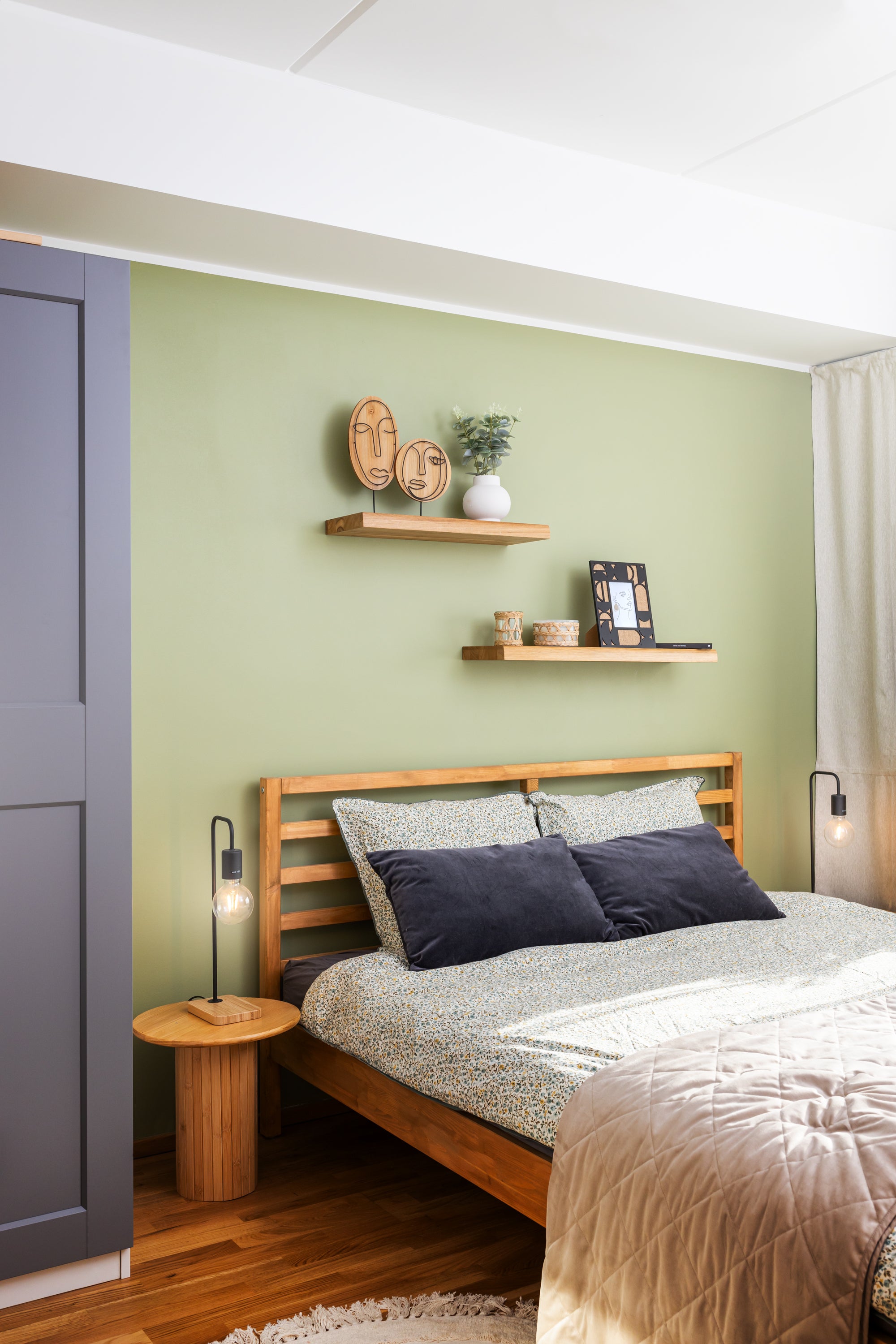 Oak floating shelves TUUL on a green bedroom wall. Bedroom has a solid wood bed and small wooden bedside tables