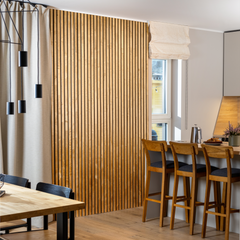 Warm toned open living room space showcasing solid wood wall panels on the wall, a solid wood dining table in the front and three bar chairs near the kitchen counter.