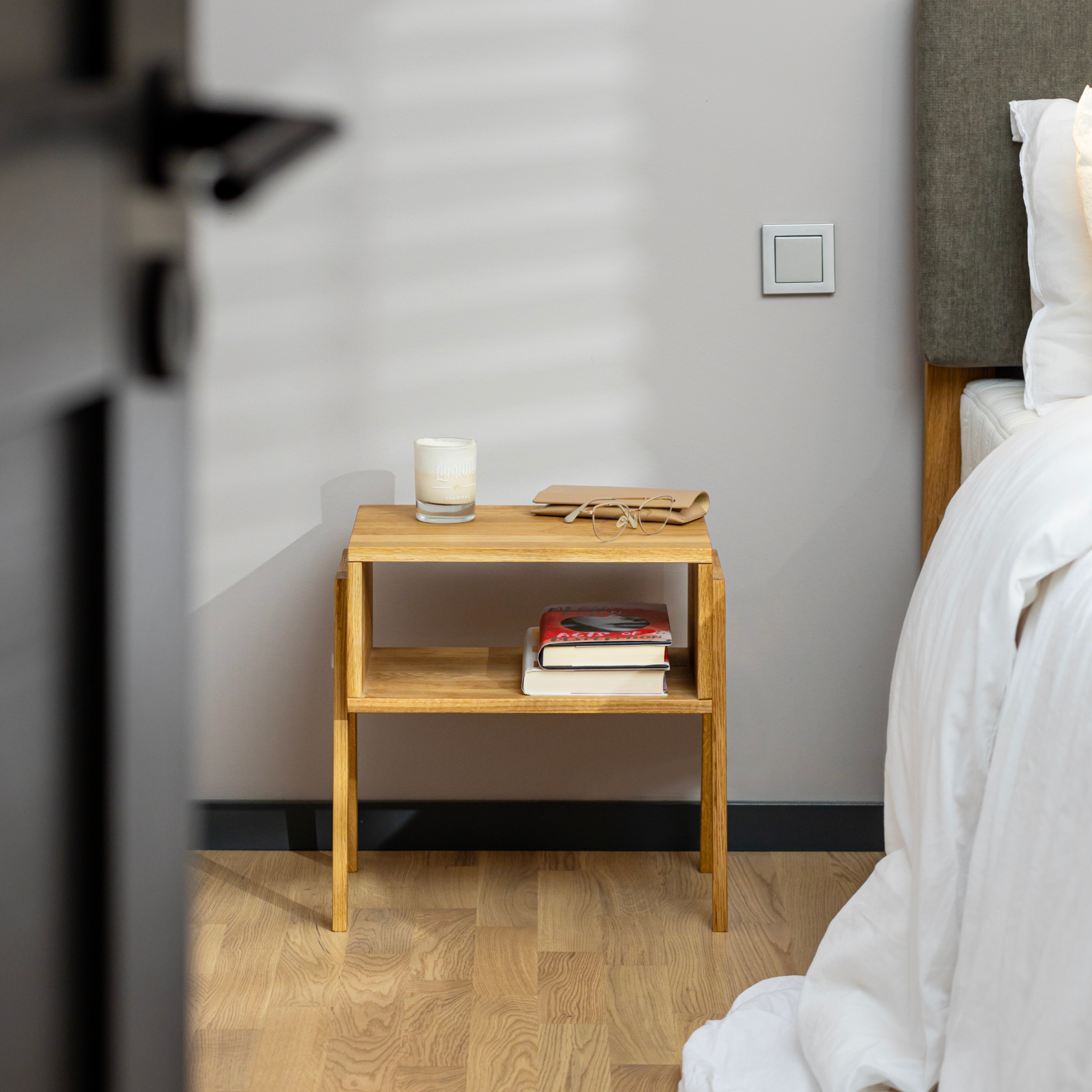 Table de chevet en bois massif KUU dans une chambre à coucher décorée avec des livres sur l'étagère inférieure et une bougie sur l'étagère supérieure.