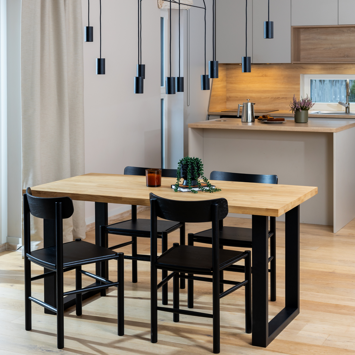 Solid oak dining table with black metal legs in an open kitchen and dining room. The table has matching black solid wood chairs. A grey kitchen is in the background