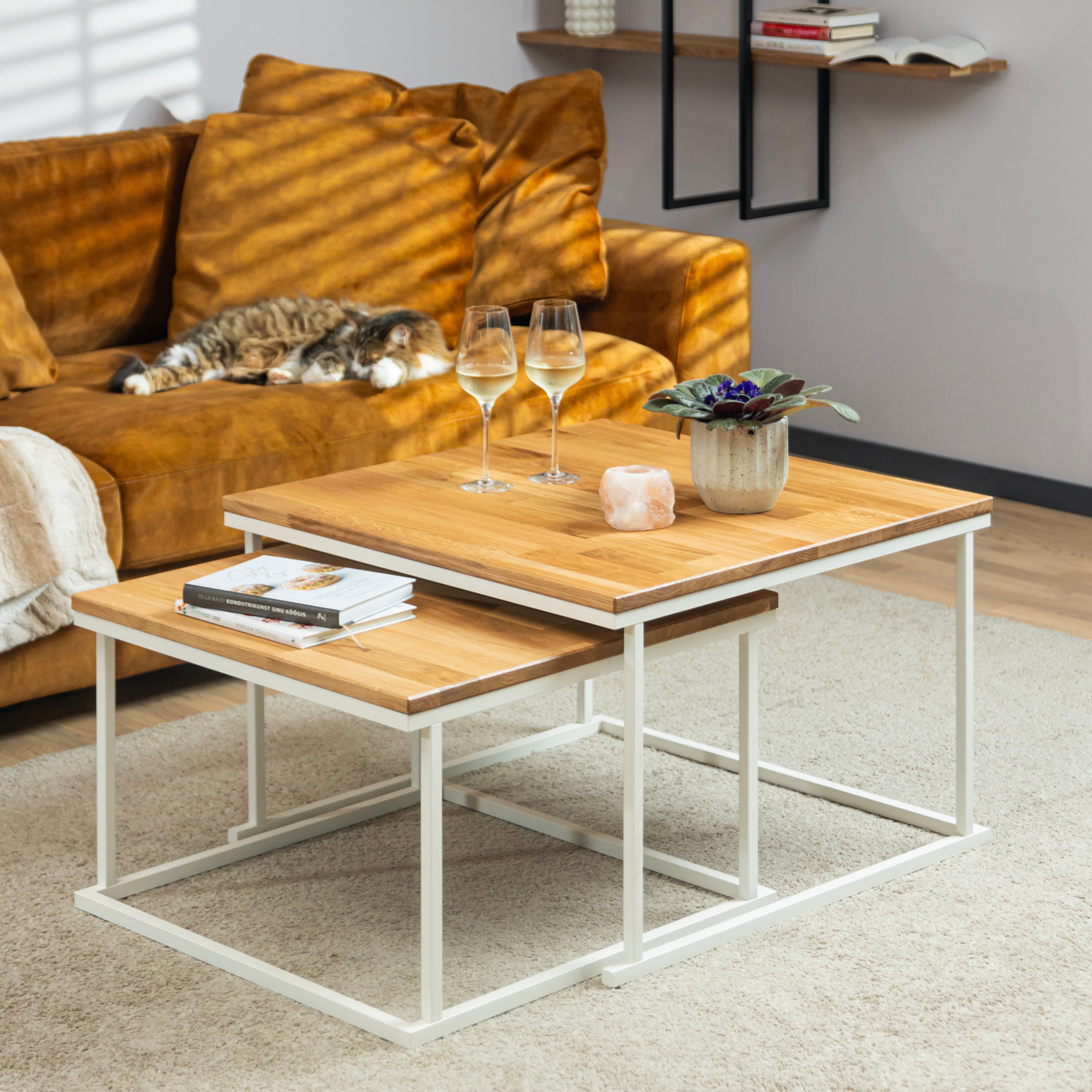 La table basse Nurmenukk en chêne avec des pieds en métal blanc est agrémentée de livres, d'une fleur, d'une bougie et de deux verres à vin pour un salon confortable.