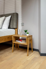 Table de nuit en bois massif KUU dans une chambre à coucher décorée avec des livres sur l'étagère inférieure et une bougie sur l'étagère supérieure.