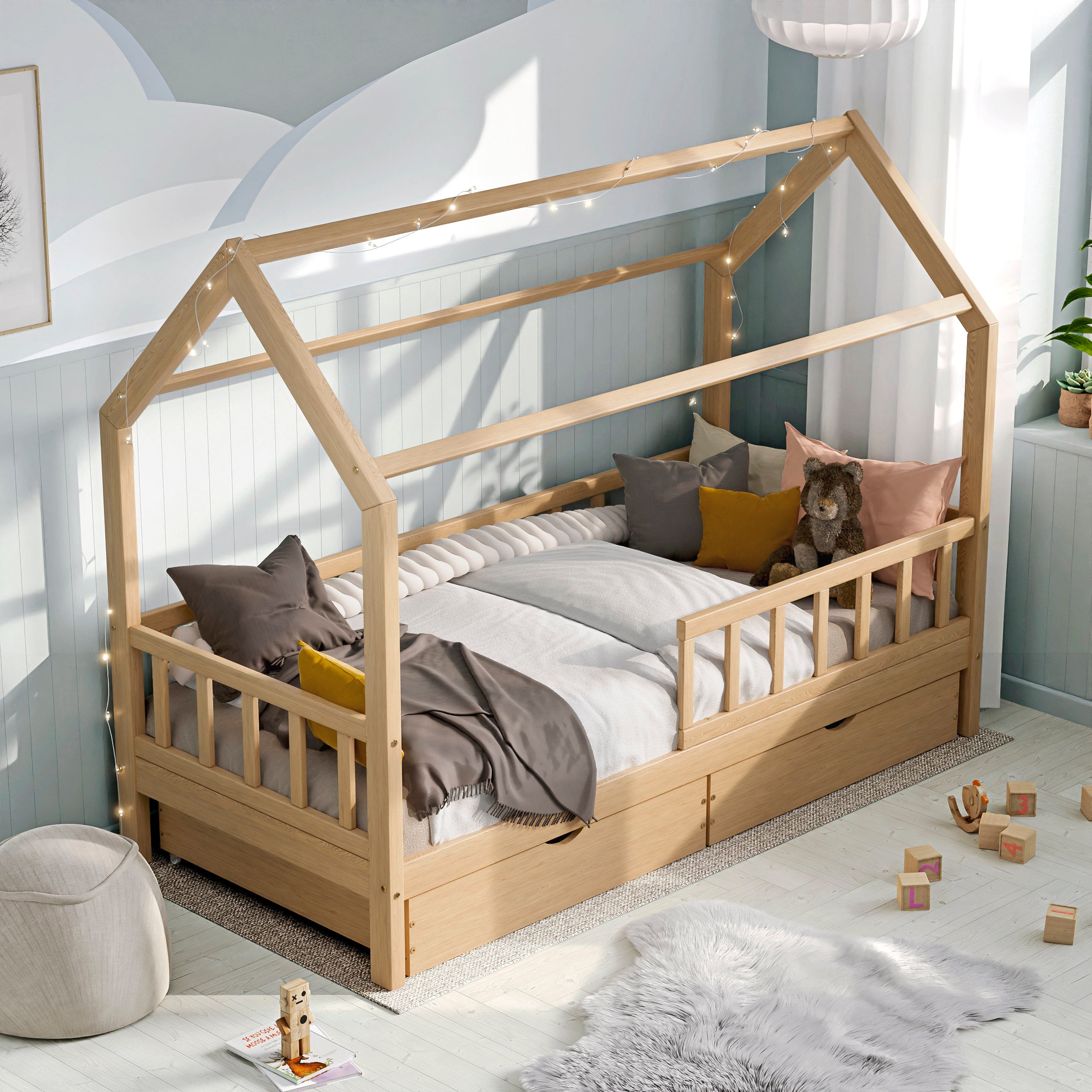 Lit d'enfant en bois naturel ÖÖBIK avec deux tiroirs et barrières dans une chambre d'enfant moderne et lumineuse décorée avec des coussins et des jouets.