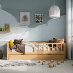 Vue de face du lit d'enfant en bois avec barrières, tiroirs et lit escamotable dans une chambre d'enfant décorée de coussins et de jouets.