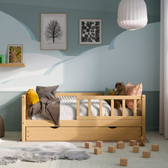 Front view of the wooden toddler bed with barriers, drawers and a pull-out bed in a kids room decorated with pillows and toys